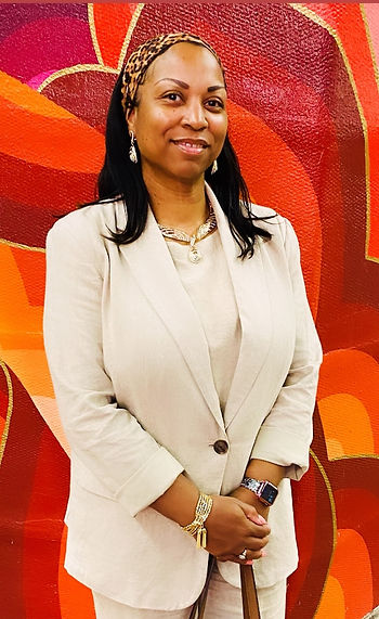 Black Woman in an eggshell pant suit, standing in front of a red wall.