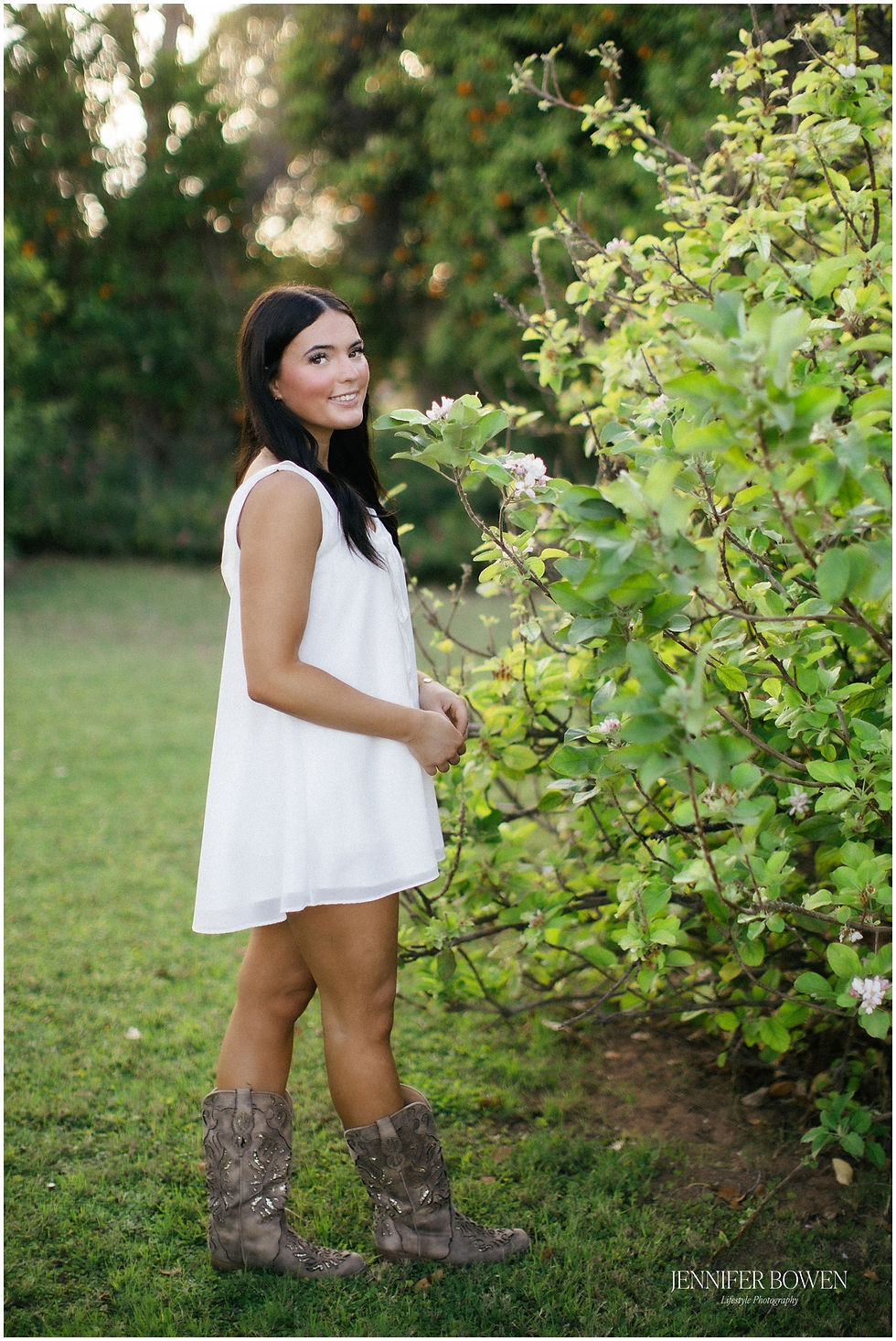 Senior photos in green location in Gilbert AZ - young woman wears white dress and brown cowboy boots for her grad portraits. Photo by Jennifer Bowen