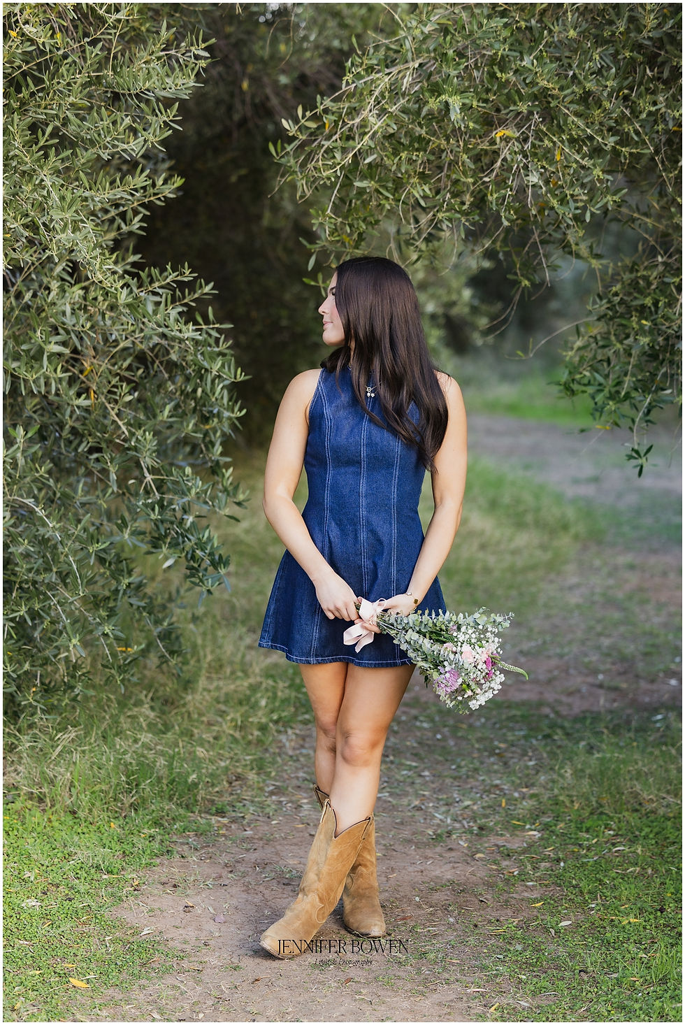 Woman in denim dress and boots holds flowers, standing on a path surrounded by green foliage, looking to her right. Peaceful mood.