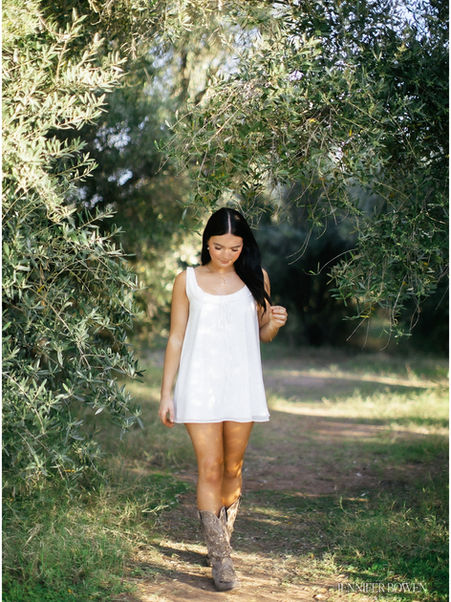 A young women walks through sunlit olive trees in a white dress and boots. Senior photos in Arizona.