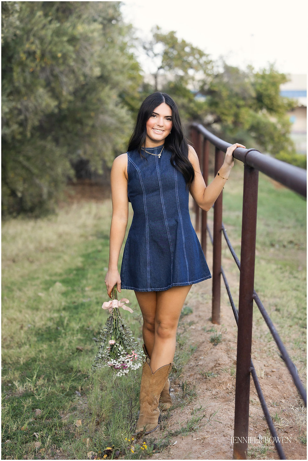 Senior photos at green location in Arizona, olive grove farm. Young woman wears a denim mini dress and brown suede cowboy boots, holding flowers. Photo by Jennifer Bowen