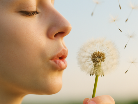 Primo piano di una donna con gli occhi chiusi che soffia su un soffione, mentre i semi volano via nel cielo; immagine luminosa, serena, simbolo di rilascio e benessere.