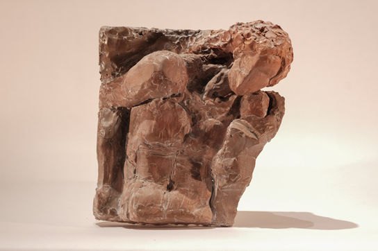 Terracotta-colored clay relief depicting two intertwined, contorted human figures emerging from a rectangular block, their forms rough and tactile.