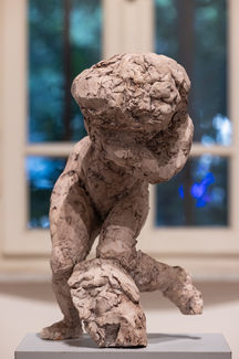 Close-up of a rough, abstract clay figure in a crouched stride, displayed on a plinth with a softly lit window backdrop.