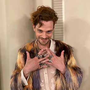 Dutch young countertenor GERBEN VAN DER WERF as Tolomeo in his dressing room of Castle Schwetzingen. A young man with dark-blond hair, a curl at his forehead, wearing a white silk shirt and a coat of feathers in brown, beige, lilac, orange and light yellow. In front of his chest, he is showing his hands with many silvers rinngs on each hand. He is smiling sarcastically - Händel- Giulio Cesare, production by Nationaltheater Mannheim