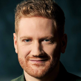 Tenor Merlin Wagner mit rotblondene Haaren, Bart und grünen Augen lächelt verschmitzt - Kamala Sankaram's opera Thumbprint at Opera Wuppertal