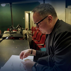 German stage-director HENDRIK MÜLLER sitting in front of a tabel and a scsore of J. Strauß "Die Fledermaus" in the Landestheater Flesnburg- Rehearsal atmosphere, middle-aged man with short hair, big black glasses, black t-Shirt, balck blazer, a watch at his wrist
