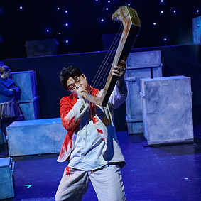 Beomjin Kim, a Korean tenor on stage with several stones and boxes in light purle, in a white-red costume trying to make music on a harp, blowing it, instead of playing. Children's opera at Badisches Staatstheater Karlsruhe: Orpheus & the magic harp