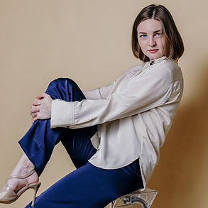 Anastasiya Taratorkina, woman begin 30th, white, blue eyes, red lipe, middle blond shoulder long hair, sitting on a chair, wearing a beige silk bluse, beige pumps dn a dark blue silk pants, hodling her red knee with both hands and looking tothe camera over her left shoulder