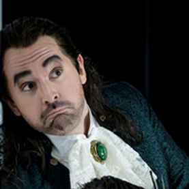 Tenor Rafael Helbig Kostka in costume: Man begin 30th,, with ablack long wigg, beart, brown eyes, wrikelnd forehead looking to the left, dressed in a blue-green baroque jacket, a white baroque shirt with a huge green-golden broche in the middle - Nationaltheater Mannheim