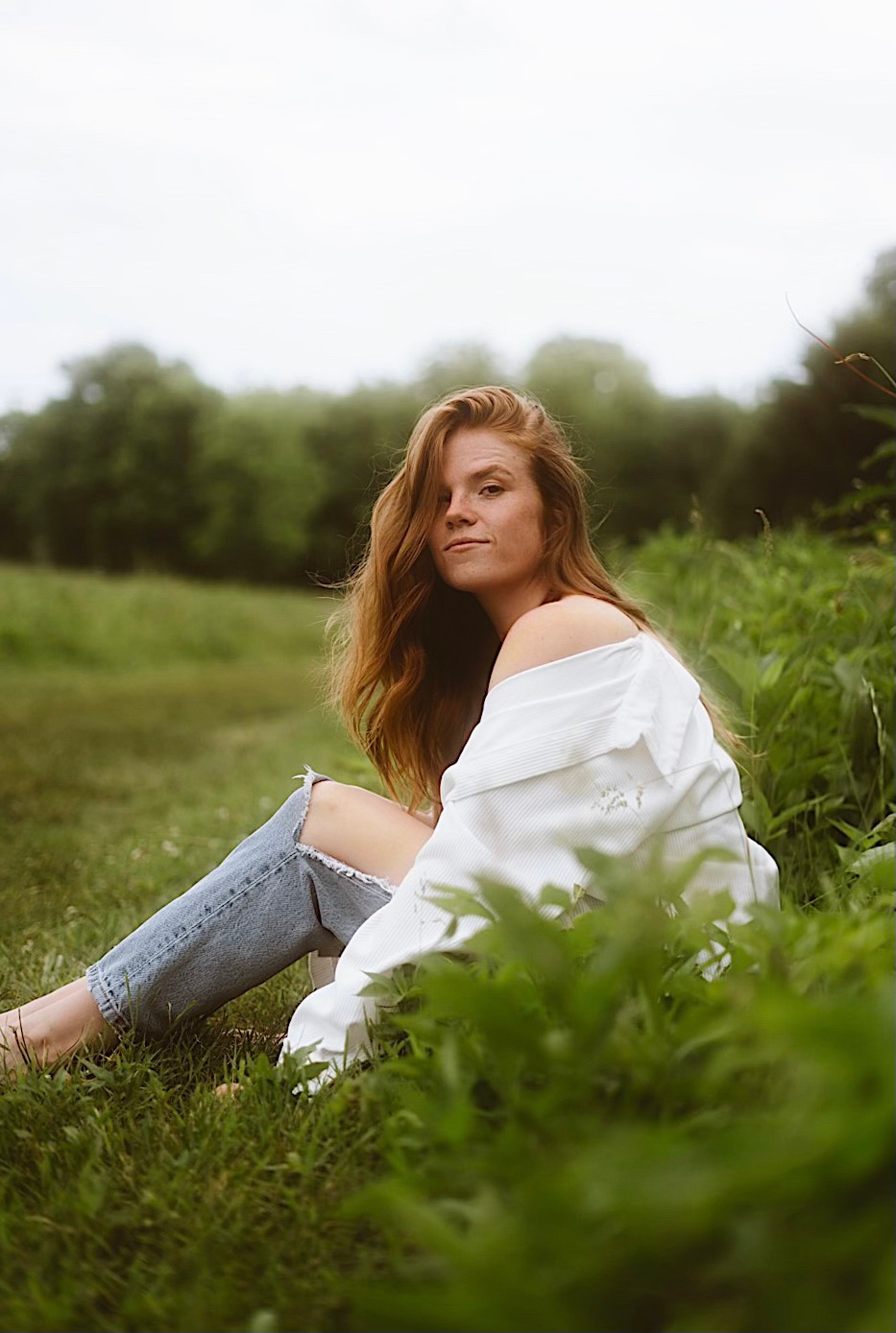 A woman sitting in a field in a white shirt and blue jeans smiling