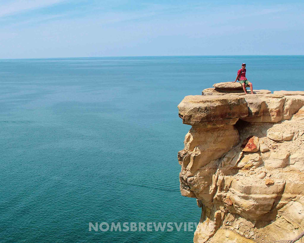 Person in red shirt sits on a rocky cliff overlooking the ocean under a clear blue sky. Text "NOMSBREWSVIEWS" is visible. Mood is adventurous.
