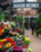 Flower shop with vibrant bouquets and cacti on display. A person with a backpack walks under a "Maison Meunier" sign. Bright, lively setting.