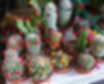 Cacti with colorful artificial flowers glued on top sit in small pots on a table. The scene is vibrant and lively.