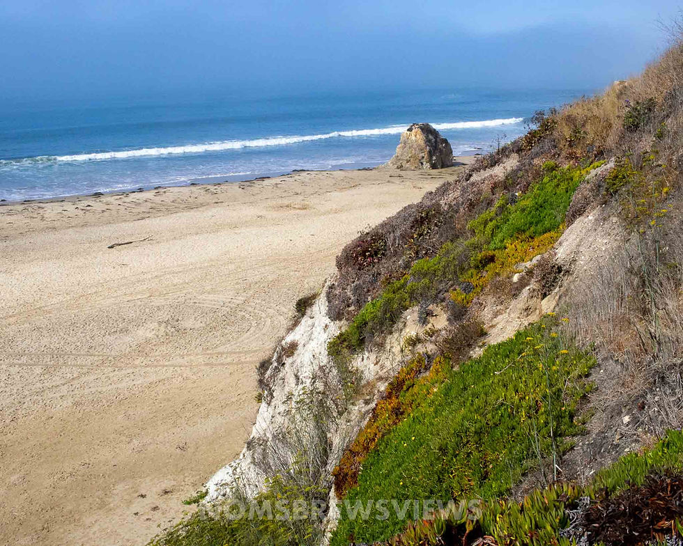 Sandy beach with a rock formation by the ocean. Green and brown vegetation cover a hillside in the foreground. Calm, sunny atmosphere.