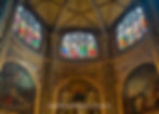 Stained glass windows in a cathedral with vibrant colors above ornate murals. Latin text lines the golden arches, creating a serene atmosphere.