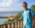 A person in a blue shirt and sunglasses smiles at a beach overlook. The beach below is crowded with umbrellas, and the sea is calm under a blue sky.