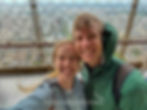 Couple smiling on Eiffel Tower observation deck, cityscape below. She's in a light jacket; he's in a green Columbia jacket. Overcast sky.