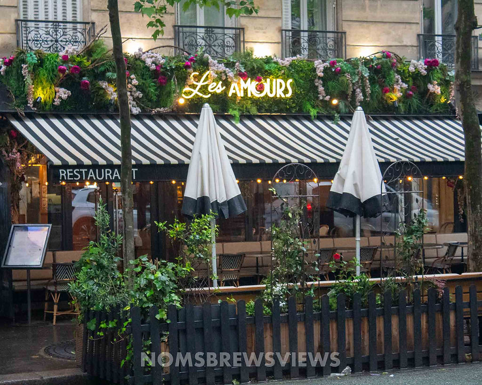 Street view of "Les Amours" restaurant with striped awning and floral decor. Closed umbrellas, greenery, and a cozy Parisian vibe.