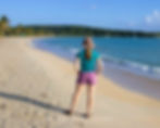 A person in a hat stands on a sandy beach, facing the ocean. Swimmers are in the water, with palm trees and a boat in the background. Bright day.