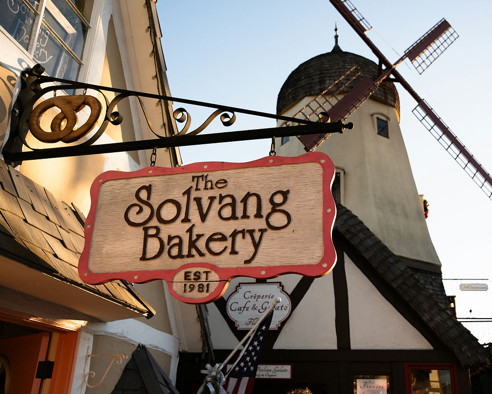 Sign reading "The Solvang Bakery" with a pretzel hangs in front of a building with a windmill in the background, under a clear sky.