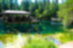 A group of people are sitting in a pavilion on a clear blue lake surrounded by lush green forest, reflecting a peaceful, sunny atmosphere.