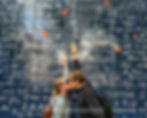 A couple kisses in front of a wall inscribed with "I love you" in various languages. The wall has a dark blue background with white text.