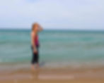 Red-haired woman stands at shoreline, hand shading eyes, facing ocean. Wearing tank top and leggings. "NOMSBREWSVIEWS" text visible in sand.