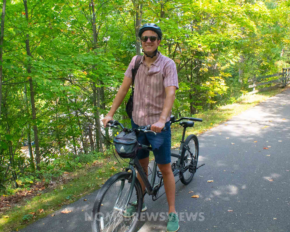 Man smiling in helmet and sunglasses, standing with a bicycle on a forested path. Green trees surround the sunny trail.