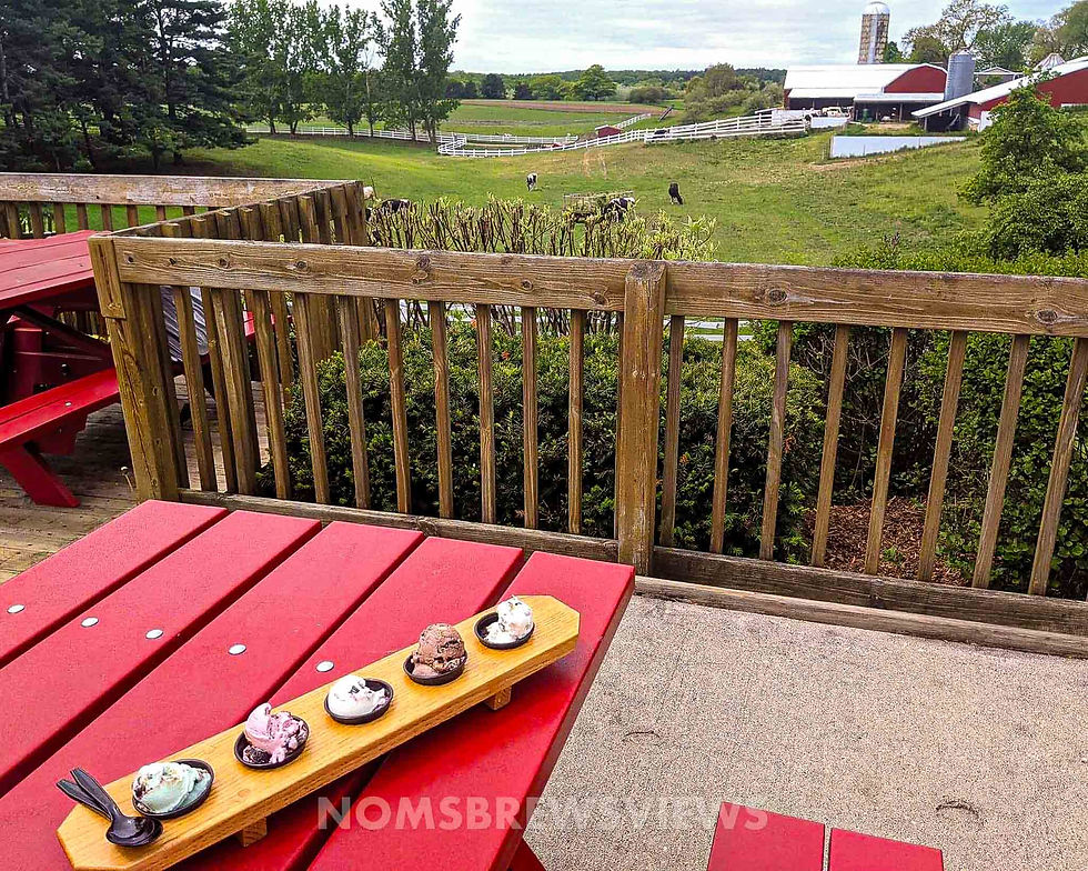 Ice cream flight on a red picnic table, overlooking a farm with red barns and grazing cows. Green fields and trees in the background.
