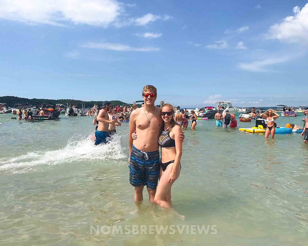 A couple poses in shallow water on a sunny day, surrounded by people and boats. The man wears red sunglasses. Text: Nomsbrewsviews.