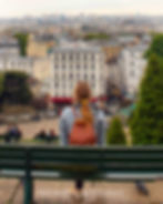 Woman with red backpack sits on a park bench overlooking a cityscape in Paris. Overcast sky, trees, and people in the background.