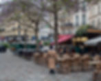 Pedestrian in a beige coat walks through an empty café courtyard with rattan chairs. Leafy trees and shops under colorful awnings line the path.