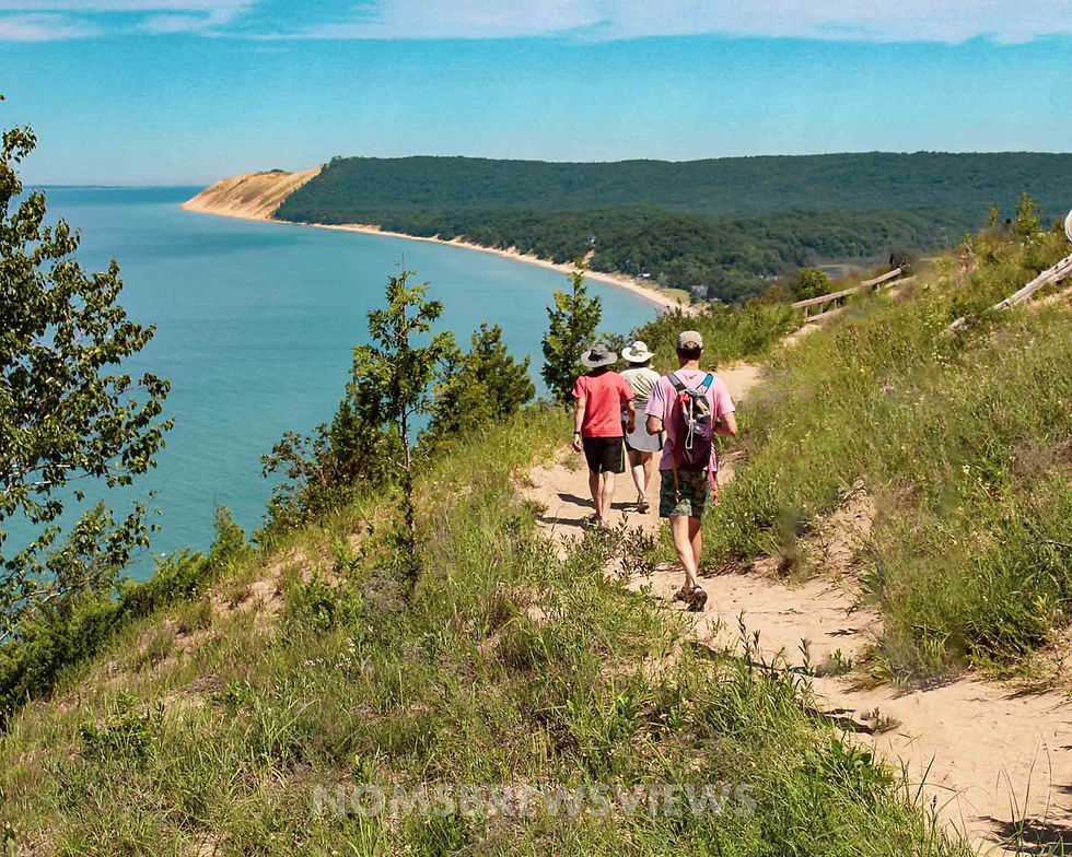 Three hikers on a sandy trail by a lush, green cliffside overlooking a blue lake. Clear sky and distant trees create a tranquil scene.