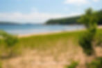 Sandy beach with green grass in foreground, calm blue water, and trees in background. Few people relax by the shore under a clear sky.