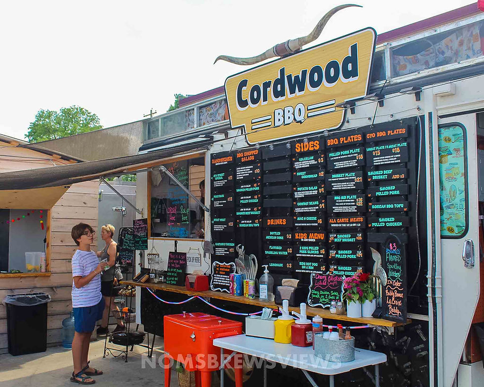 Food truck with a "Cordwood BBQ" sign, colorful menu, and condiment table. Two people standing nearby. Bright, outdoor setting.