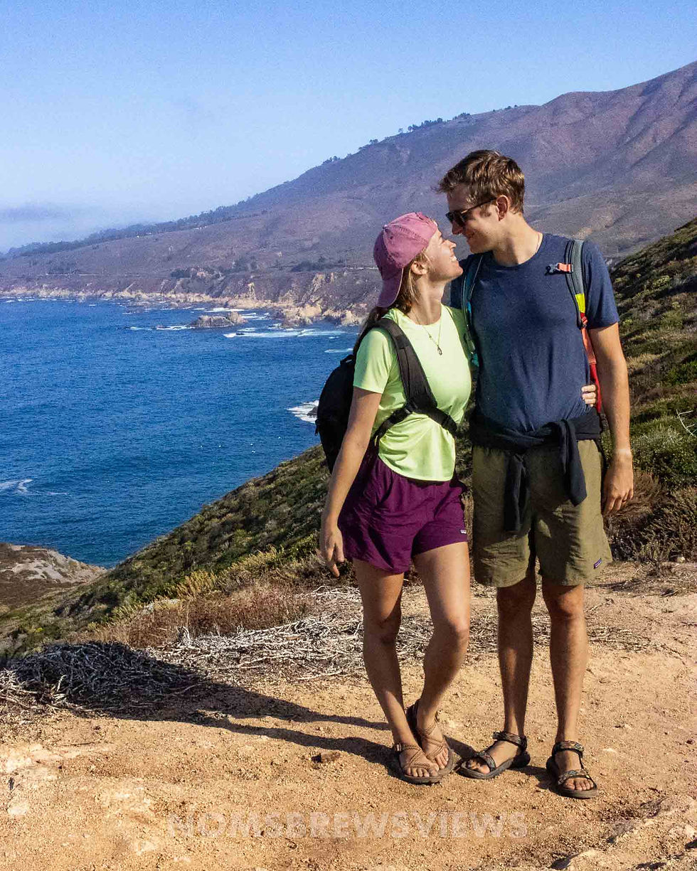 A couple with backpacks embraces on a coastal trail. The ocean and mountains are in the background, under a clear blue sky. They look happy.