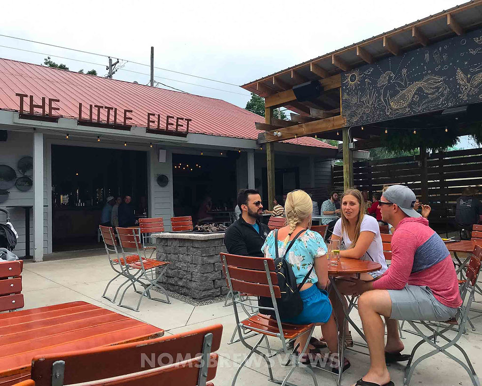 Four people seated outdoors at The Little Fleet, chatting around a table. Red chairs, mural with mermaid, casual vibe. Overcast sky.