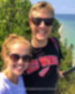 A smiling couple wearing sunglasses takes a selfie at a scenic lakeside lookout. The man wears a "RED WINGS" shirt; lush greenery surrounds them.