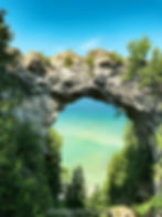 Rock arch frames turquoise lake, surrounded by lush green trees. Clear blue sky above. Calm and serene scene. Text: "nomsbrewsviews.com".