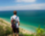 A person with a backpack stands on a grassy cliff, overlooking a vast, turquoise ocean under a partly cloudy sky. The mood is serene.