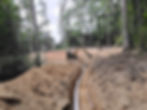 White pipe laid in a deep trench leading toward a compact excavator in a wooded area.