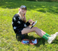 Smiling girl in star sweater sketching on a pad in green grass.