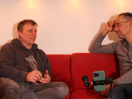Clemens Tewinkel (links) und Eddi Hüneke (rechts) sitzen gemeinsam auf einer roten Couch, während Eddi zu Clemens rüber schaut und ihm zuhört