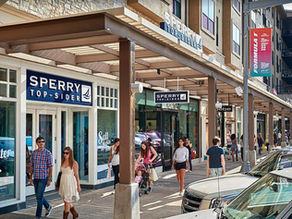 People walk along a shopping street with Sperry stores, colorful banners, and parked cars. Sunny day, casual atmosphere.