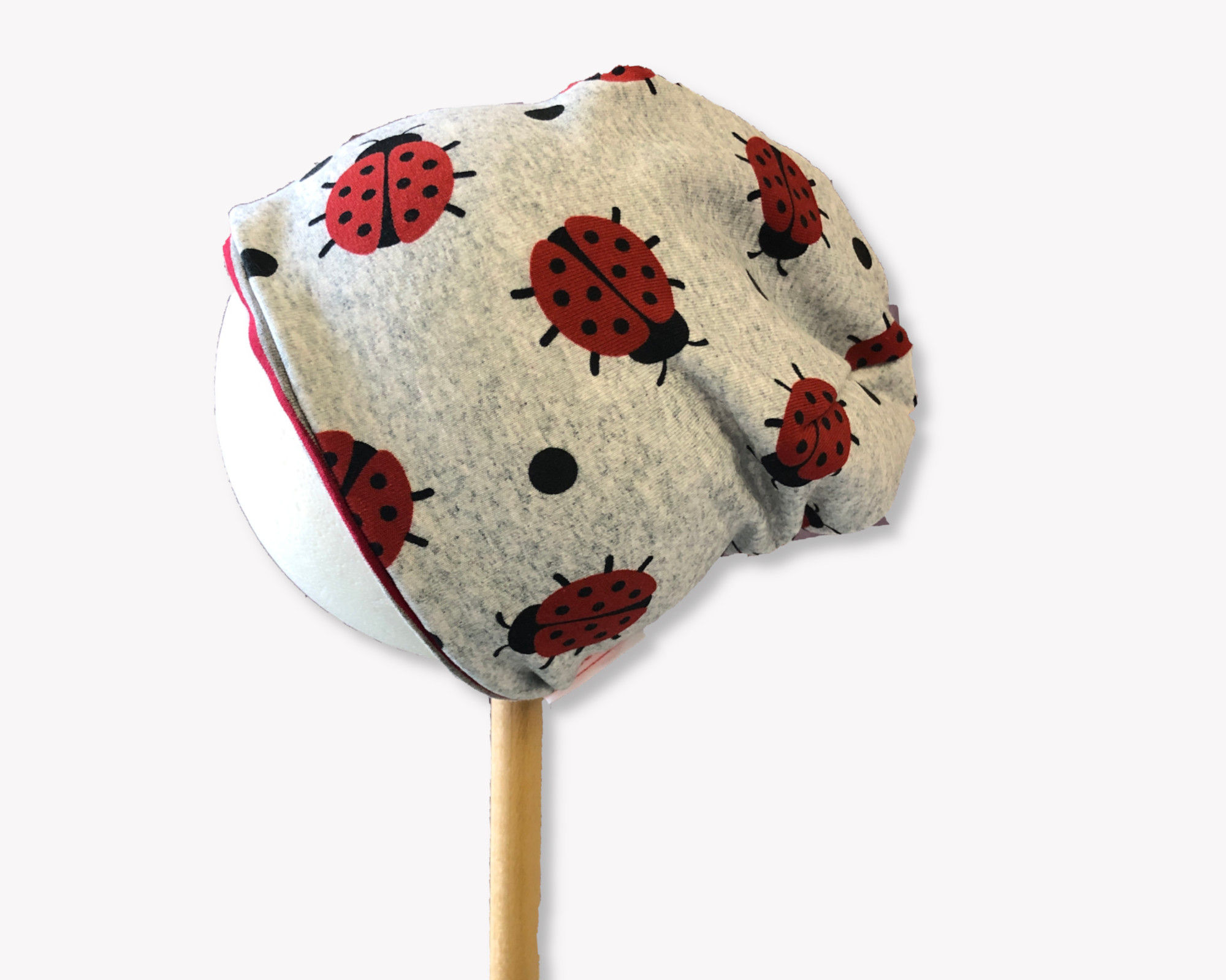 Graue Mütze mit roten Marienkäfern, sitzt auf einem Holzstab, schlichtes Design.