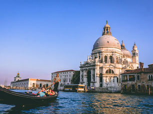 Gondola with passengers on Venice canal, passing a grand domed building at sunset. Clear blue sky and tranquil water set a serene mood.