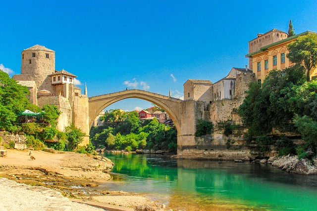 Stone bridge arches over a turquoise river in Mostar, Bosnia and Herzegovina. Sunlit buildings and lush greenery frame the scene. Clear blue sky.