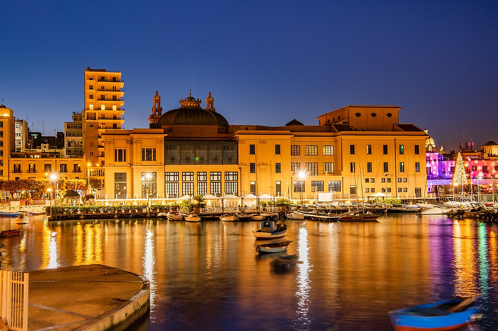 Illuminated building by a calm harbor at dusk, reflecting warm golden lights on water. Boats are docked, and a lit Christmas tree is visible.