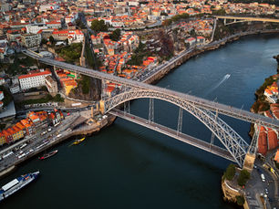 Aerial view of Porto’s Dom Luís I Bridge over the Douro River, with red-roofed city blocks, boats, and cliffs below.